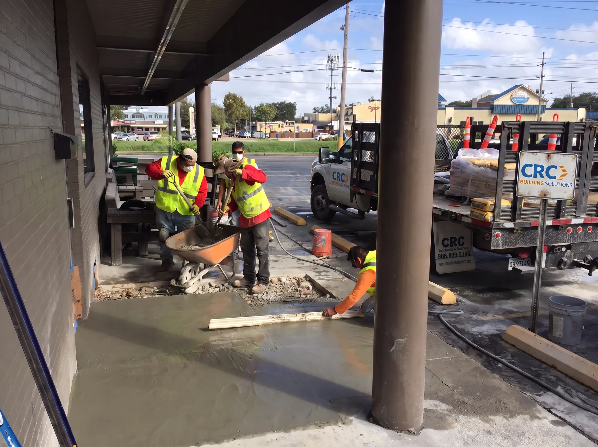 CRC Building Solutions concrete crew in hi-vis vests screeding fresh flatwork under a storefront canopy beside a CRC Building Solutions service truck in Gulfport, Louisiana