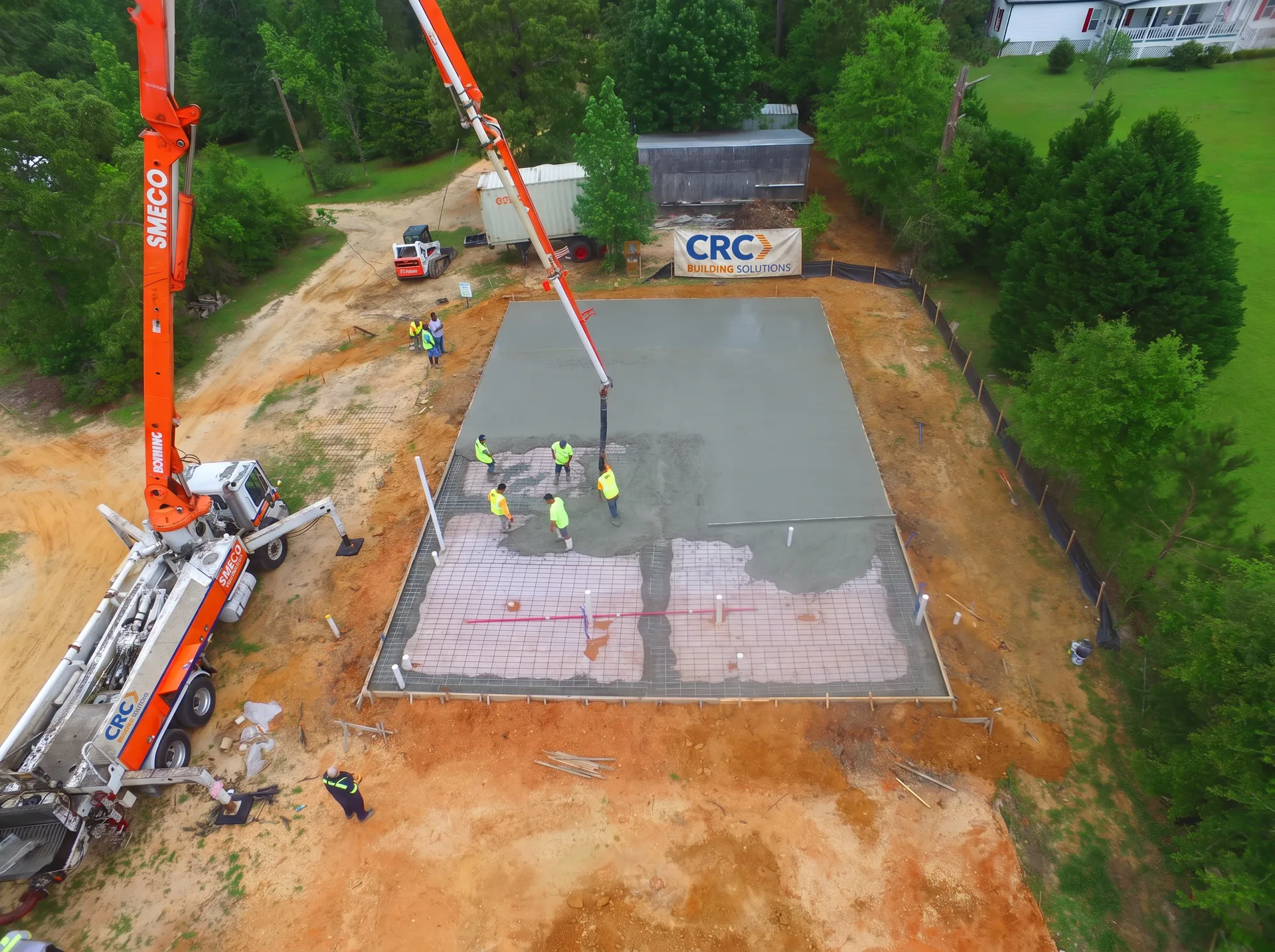 Aerial view of a CRC Building Solutions concrete crew in hi-vis vests placing a cast-in-place slab from a boom pump truck, with a CRC Building Solutions banner on the site fence