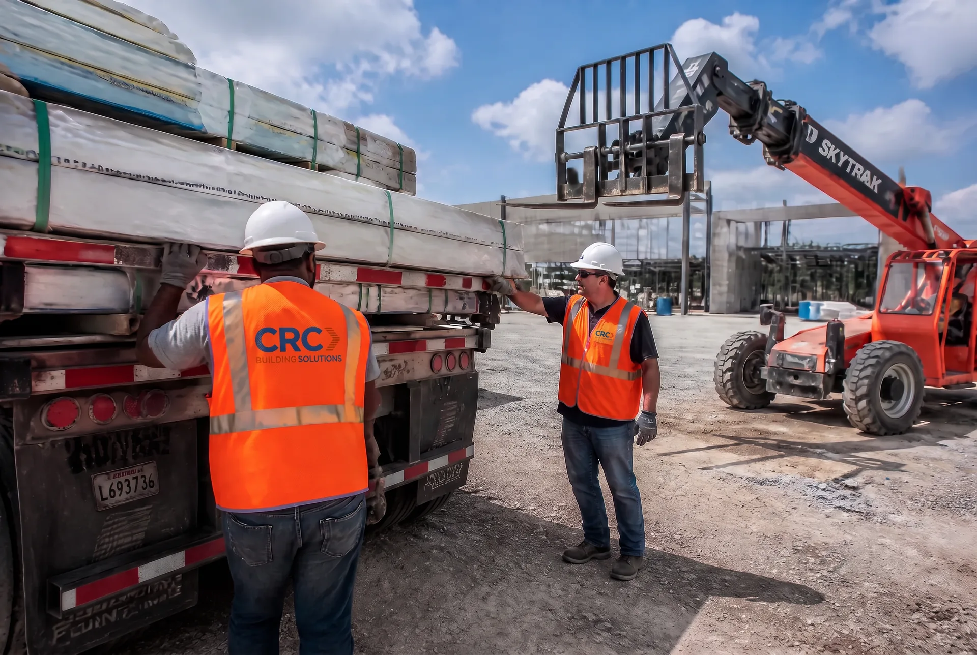 Two CRC Building Solutions crew members in CRC-branded hi-vis vests guiding banded construction materials off a flatbed trailer while a telehandler stages the load at a commercial jobsite