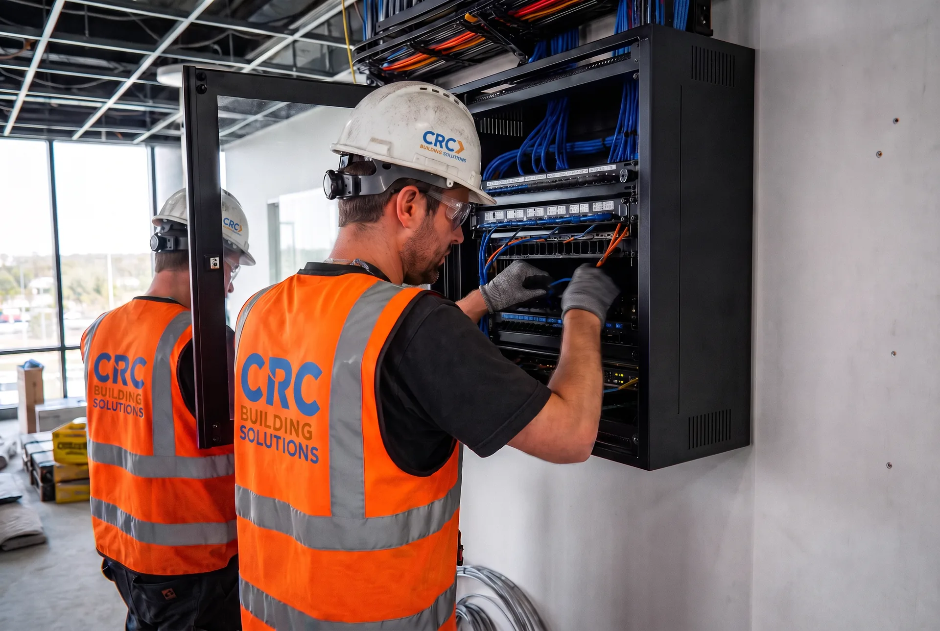 Two CRC Building Solutions low-voltage technicians in CRC-branded hi-vis vests terminating Cat6 patch cabling at a wall-mounted network rack
