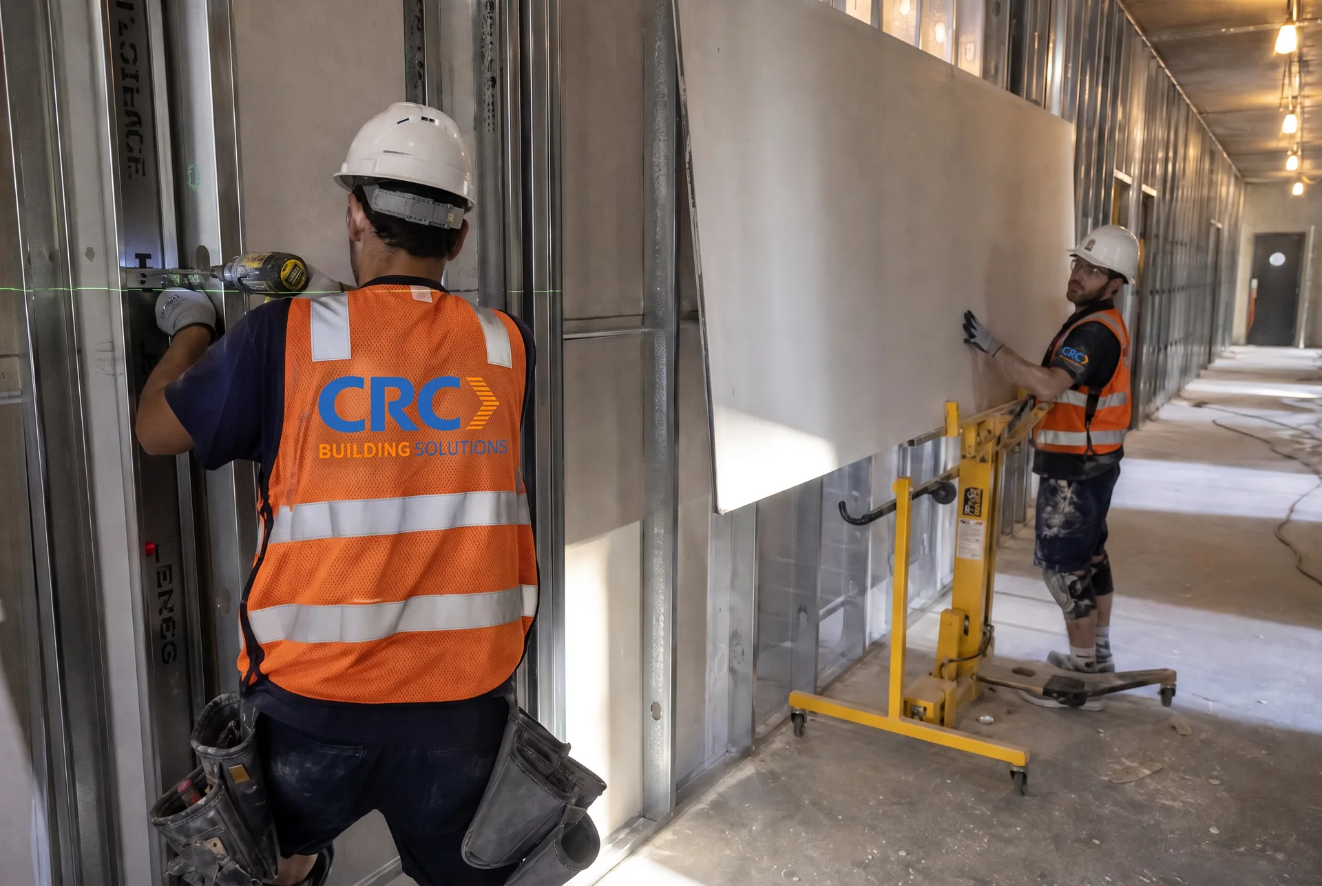 CRC Building Solutions carpenter in a CRC-branded hi-vis vest plumbing metal stud framing while a second crew member sets a gypsum board panel on a drywall lift in a commercial corridor