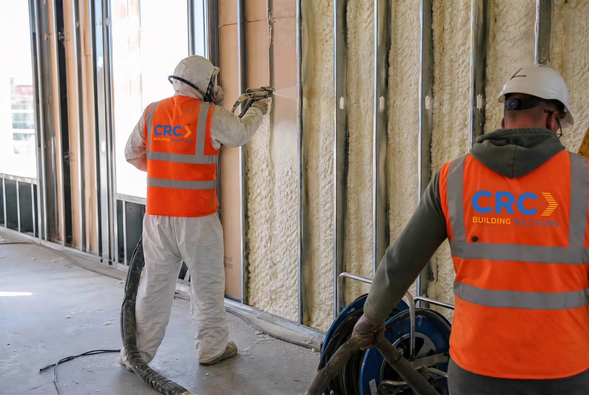 CRC Building Solutions insulation applicator in a hooded suit and CRC-branded vest spraying closed-cell foam into steel-stud wall cavities in Gulfport, Louisiana