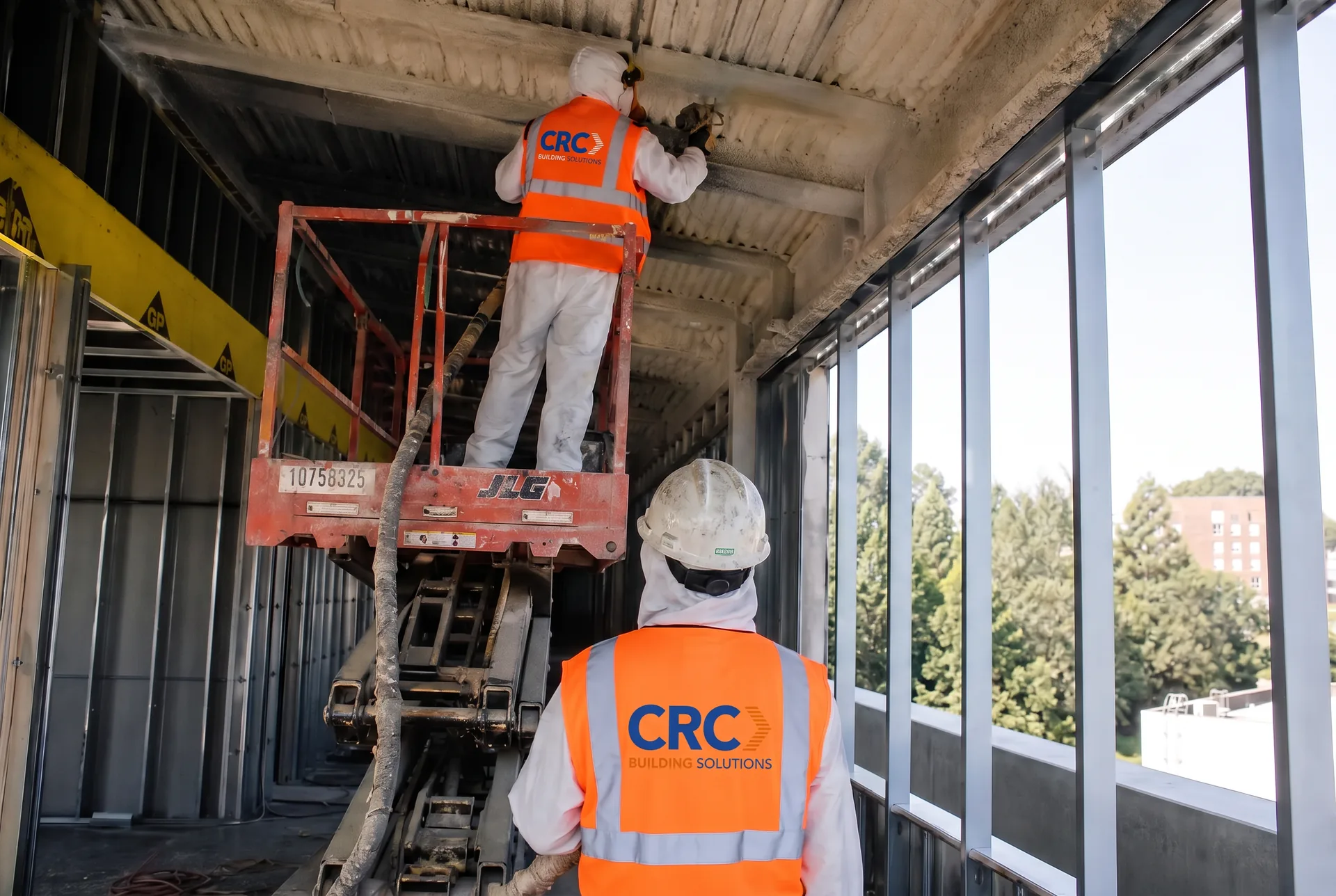 CRC Building Solutions insulation crew in CRC-branded hi-vis vests spraying closed-cell foam onto an underside deck from a scissor lift during a commercial renovation